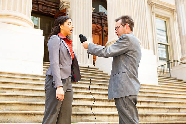 A man with a microphone interviewing a professionally dressed woman on the steps of a building. She could be a politician.